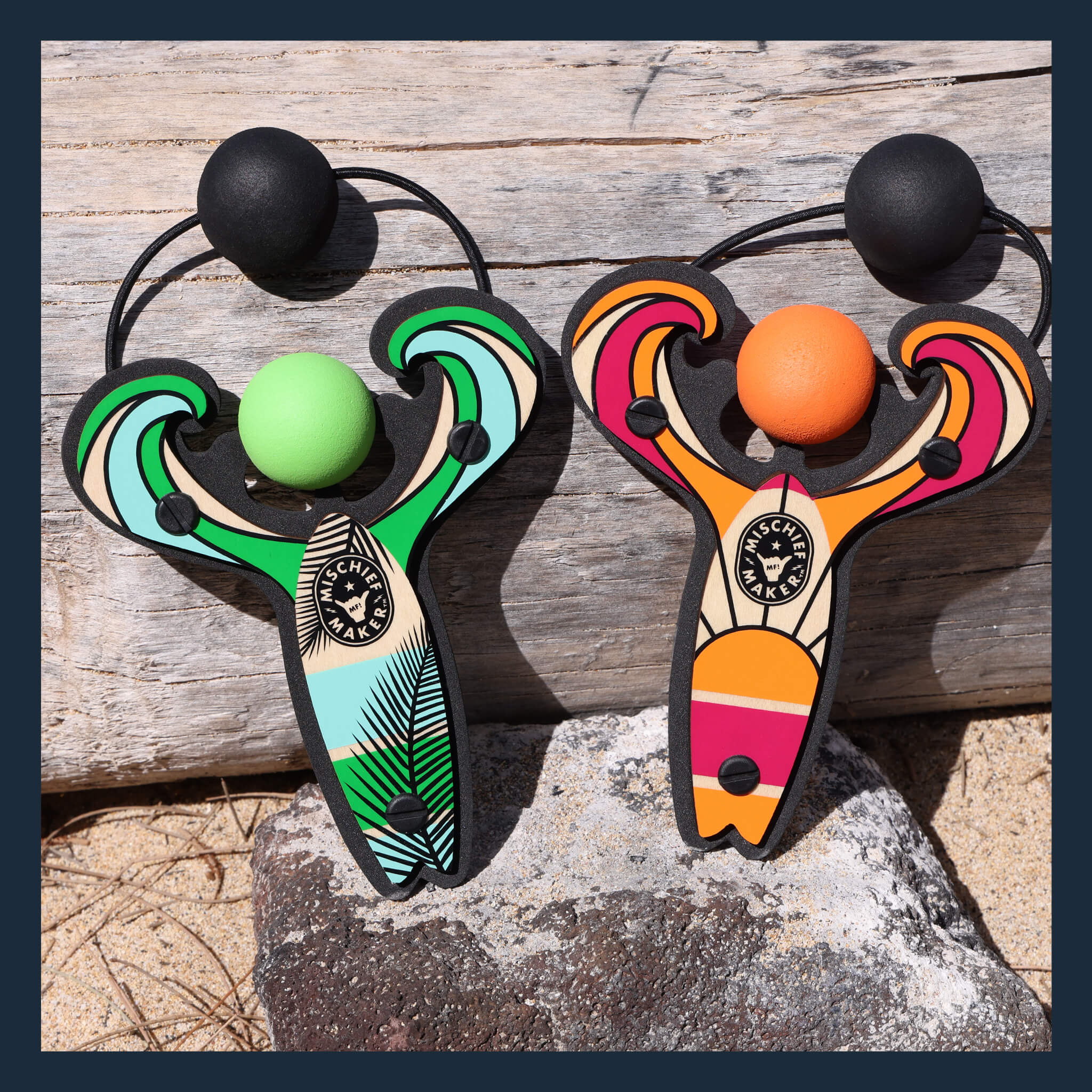 Green and Orange Surf’s Up toy slingshots in the sand on the beach. Mischief Maker by Mighty Fun!