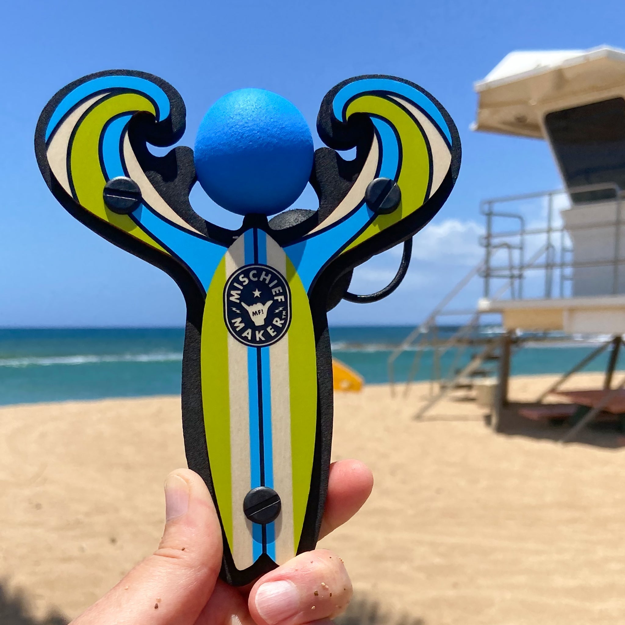 Blue Surf’s Up toy slingshot hand held by the ocean. Mischief Maker by Mighty Fun!