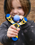 Blue classic wooden Mischief Maker kids toy slingshot being shot by 6 year old girl. By Mighty Fun!