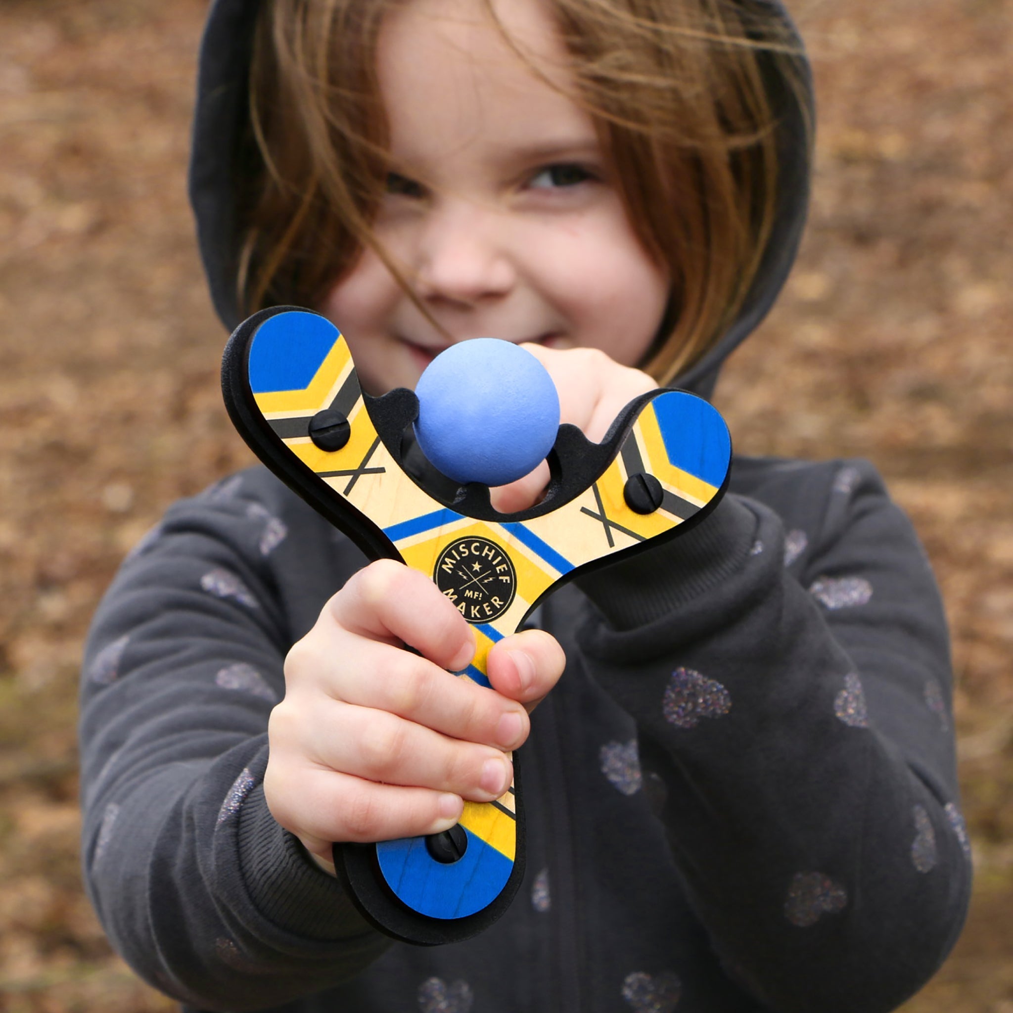 Blue classic wooden Mischief Maker kids toy slingshot being shot by 6 year old girl. By Mighty Fun!